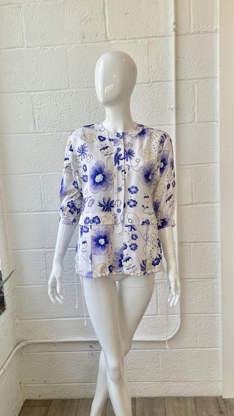 May include: White blouse with a blue floral pattern. The blouse has a round collar, three-quarter sleeves, and a button-down front. The floral design features large and small blue flowers with yellow accents.