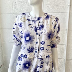 May include: White floral print top with large blue and yellow flowers. The top has a round collar, short sleeves, and two front pockets. It is buttoned down the front and has a drawstring at the waist.