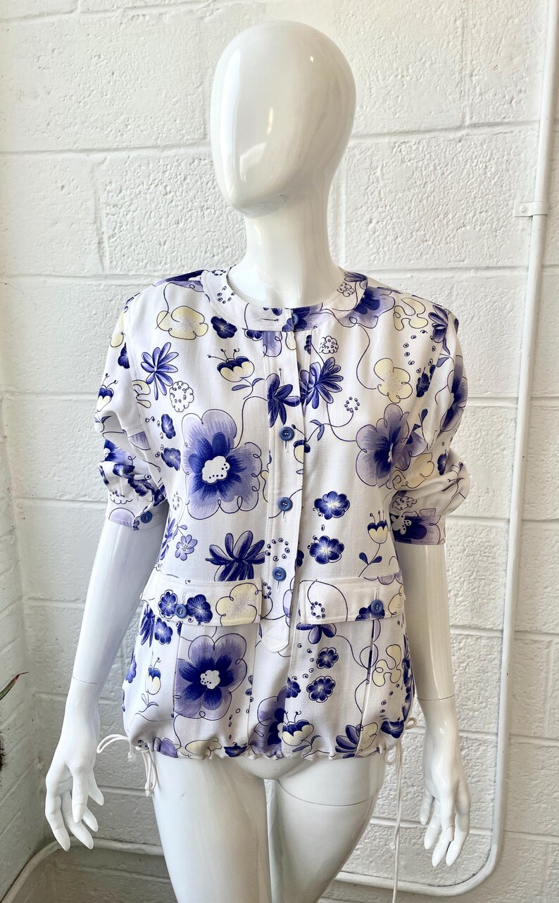 May include: White floral print top with large blue and yellow flowers. The top has a round collar, short sleeves, and two front pockets. It is buttoned down the front and has a drawstring at the waist.