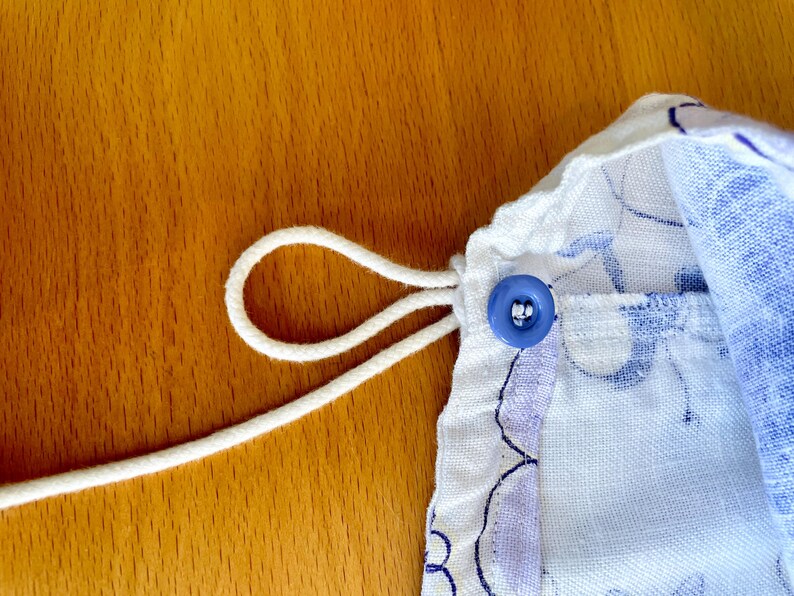May include: Close-up of a drawstring bag with a blue floral pattern. A white drawstring is looped through the fabric and secured with a blue button. The bag is on a wooden surface.