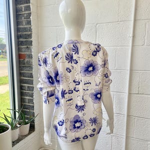 May include: A white blouse with a blue floral pattern is displayed on a mannequin. The blouse features long sleeves and a button-down front. The floral design includes large and small flowers with a delicate, feminine aesthetic.