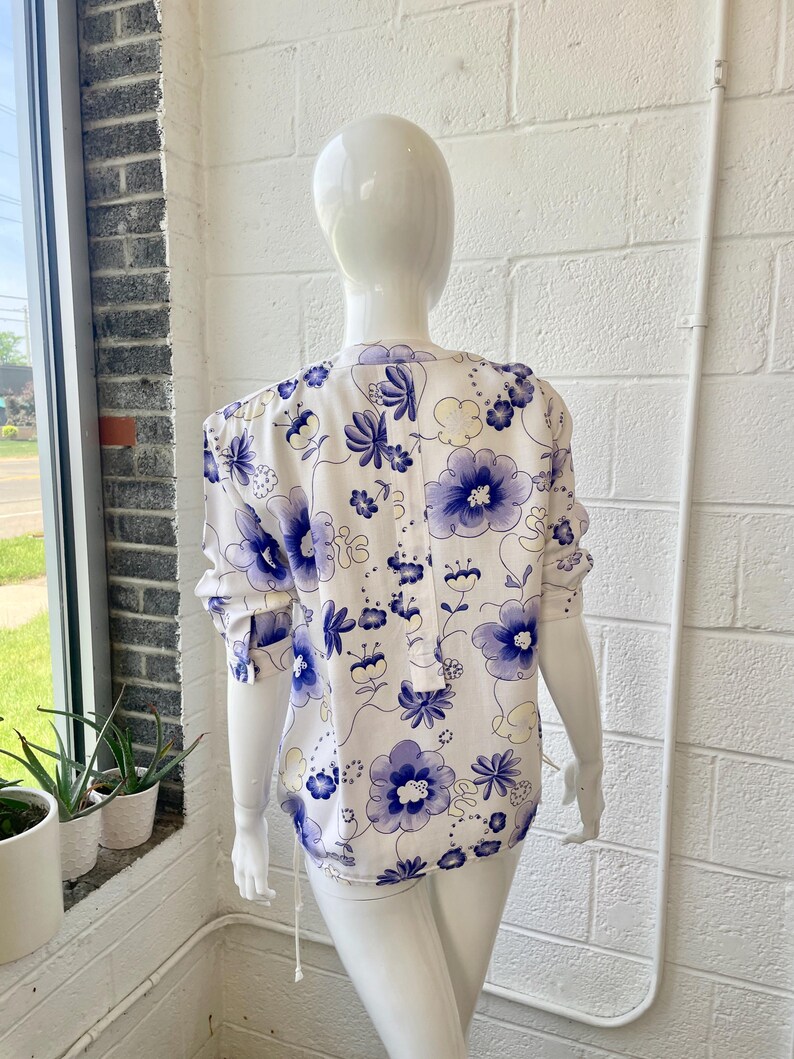 May include: A white blouse with a blue floral pattern is displayed on a mannequin. The blouse features long sleeves and a button-down front. The floral design includes large and small flowers with a delicate, feminine aesthetic.