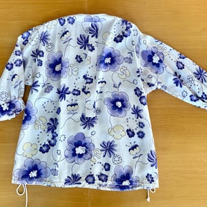 May include: White floral print top with blue flowers and green leaves. The top has long sleeves with buttoned cuffs and a drawstring at the bottom. The floral pattern is repeated across the entire top.