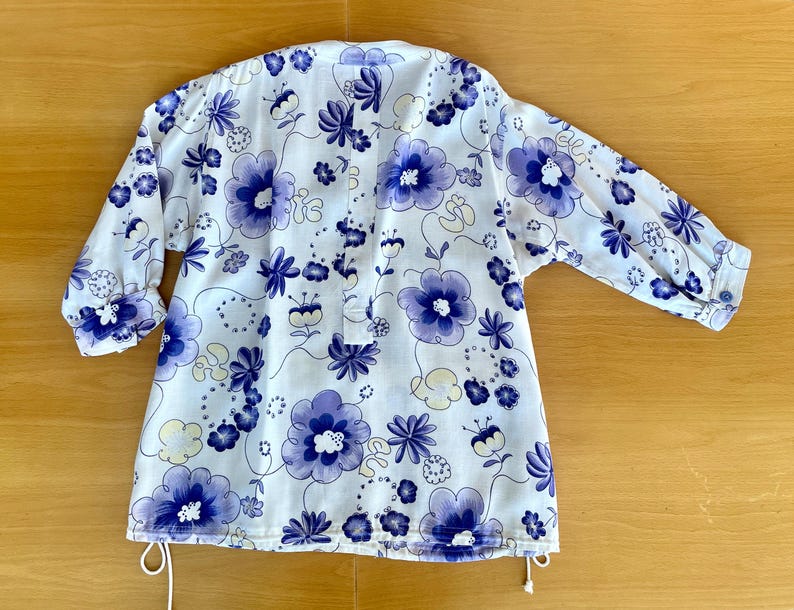 May include: White floral print top with blue flowers and green leaves. The top has long sleeves with buttoned cuffs and a drawstring at the bottom. The floral pattern is repeated across the entire top.