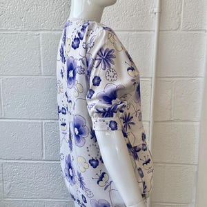 May include: A white blouse with a blue floral pattern is displayed on a mannequin. The blouse features a round neckline and long sleeves with buttoned cuffs. The floral design includes large and small blue flowers with yellow accents.