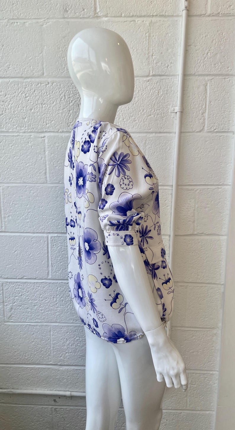 May include: A white blouse with a blue floral pattern is displayed on a mannequin. The blouse features a round neckline and long sleeves with buttoned cuffs. The floral design includes large and small blue flowers with yellow accents.