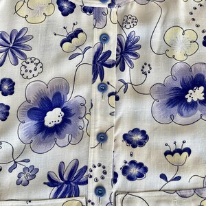 May include: White button-up shirt with a floral pattern in shades of blue and pale yellow. The shirt features a round collar, blue buttons, and two front pockets. The floral design includes large, stylized flowers and smaller floral accents.