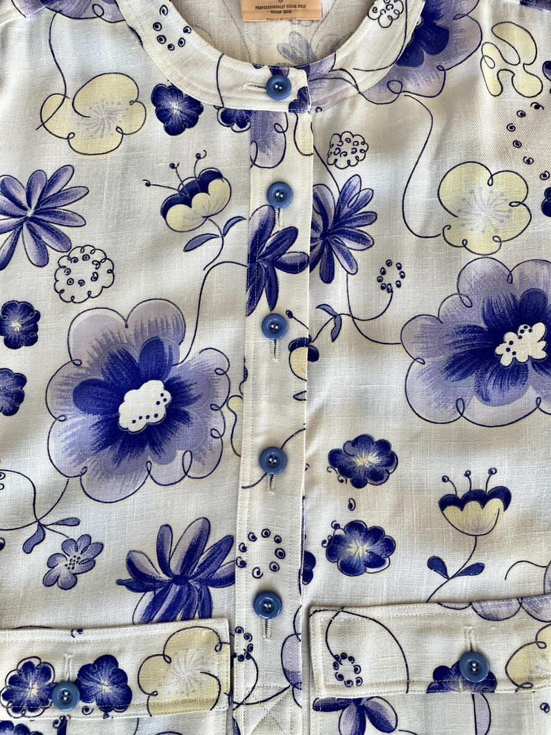 May include: White button-up shirt with a floral pattern in shades of blue and pale yellow. The shirt features a round collar, blue buttons, and two front pockets. The floral design includes large, stylized flowers and smaller floral accents.