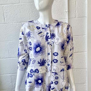 May include: White floral print jacket and matching shorts set. The set features a pattern of large blue flowers and smaller blue and yellow floral accents. The jacket has a round collar, three-quarter sleeves, and blue buttons. The shorts have pockets and a drawstring.