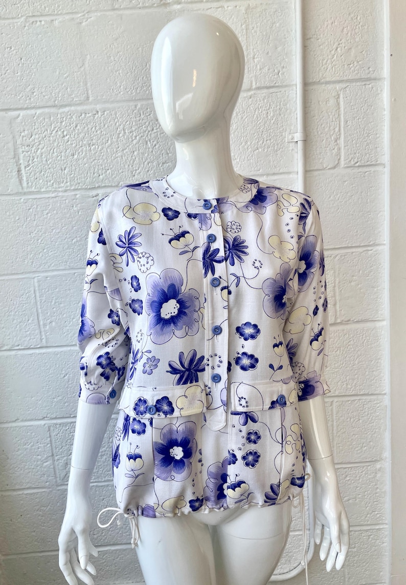 May include: White floral print jacket and matching shorts set. The set features a pattern of large blue flowers and smaller blue and yellow floral accents. The jacket has a round collar, three-quarter sleeves, and blue buttons. The shorts have pockets and a drawstring.