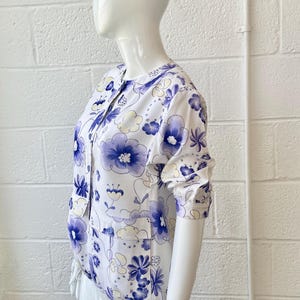 May include: White blouse with a blue floral pattern. The blouse has a round collar, button-up front, and long sleeves. The floral design features various blue flower shapes and light yellow accents.