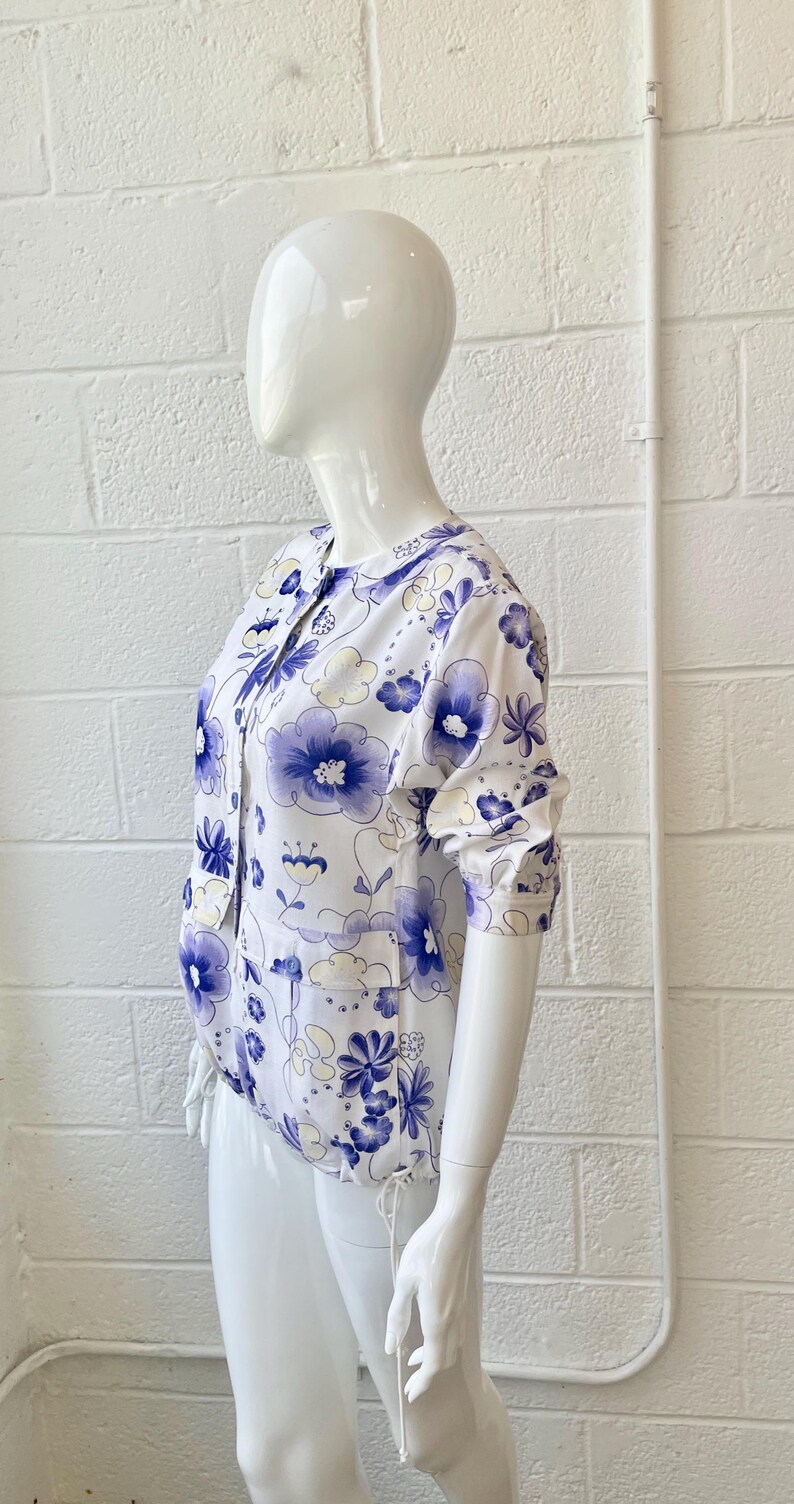 May include: White blouse with a blue floral pattern. The blouse has a round collar, button-up front, and long sleeves. The floral design features various blue flower shapes and light yellow accents.