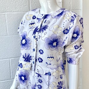 May include: White floral print blouse with short sleeves and a button-up front. The blouse features a pattern of blue and yellow flowers. The garment is displayed on a mannequin. The blouse has a drawstring at the waist.