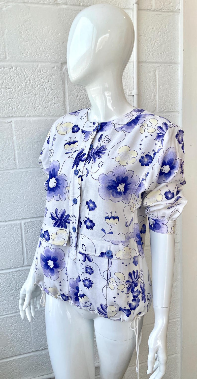 May include: White floral print blouse with short sleeves and a button-up front. The blouse features a pattern of blue and yellow flowers. The garment is displayed on a mannequin. The blouse has a drawstring at the waist.