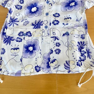 May include: White button-up shirt with a floral pattern in shades of blue and purple. The shirt features two front pockets, blue buttons, and drawstrings at the bottom hem. The floral design includes large and small flower motifs.