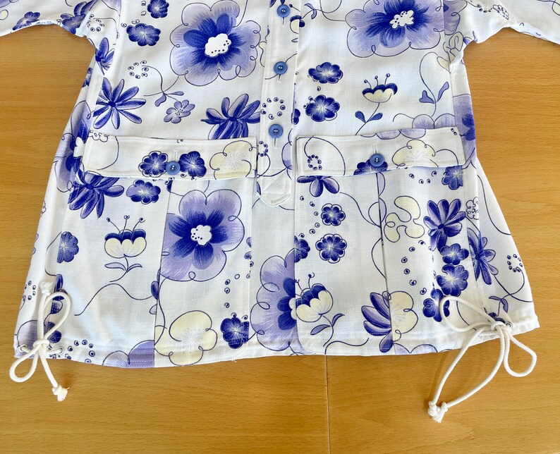 May include: White button-up shirt with a floral pattern in shades of blue and purple. The shirt features two front pockets, blue buttons, and drawstrings at the bottom hem. The floral design includes large and small flower motifs.