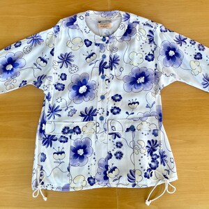 May include: White floral print jacket with blue and purple flowers. The jacket has a round collar, long sleeves, and two front pockets. It features a button-up front and drawstring details at the bottom. The floral pattern is repeated throughout the jacket.