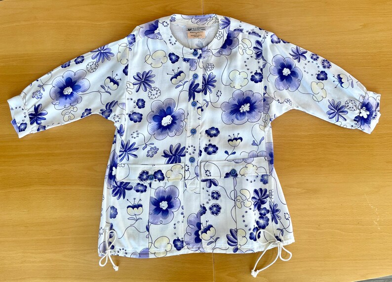 May include: White floral print jacket with blue and purple flowers. The jacket has a round collar, long sleeves, and two front pockets. It features a button-up front and drawstring details at the bottom. The floral pattern is repeated throughout the jacket.