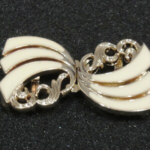 May include: A vintage bow-shaped brooch with a gold-tone metal frame and cream-colored enamel detailing. The brooch features a layered design with decorative scrollwork, creating an elegant accessory.