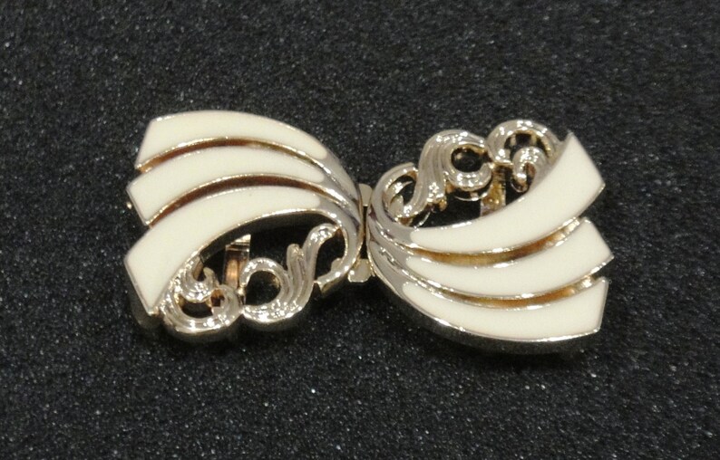 May include: A vintage bow-shaped brooch with a gold-tone metal frame and cream-colored enamel detailing. The brooch features a layered design with decorative scrollwork, creating an elegant accessory.