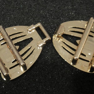 May include: Two gold-colored metal belt buckles. Each buckle has a decorative, fan-shaped design with cut-out details and two parallel bars for the belt to pass through. The buckles are on a dark background.