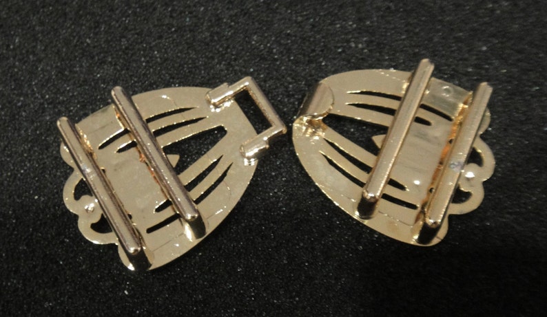 May include: Two gold-colored metal belt buckles. Each buckle has a decorative, fan-shaped design with cut-out details and two parallel bars for the belt to pass through. The buckles are on a dark background.