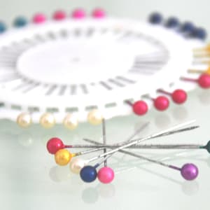 May include: A white pin wheel with a variety of colorful pins. The pins have round, pearl-like heads in shades of red, yellow, blue, green, and purple. Some pins are scattered around the wheel.