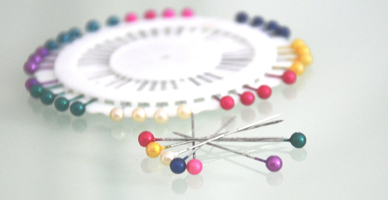 May include: A white pin wheel with a variety of colorful pins. The pins have round, pearl-like heads in shades of red, yellow, blue, green, and purple. Some pins are scattered around the wheel.