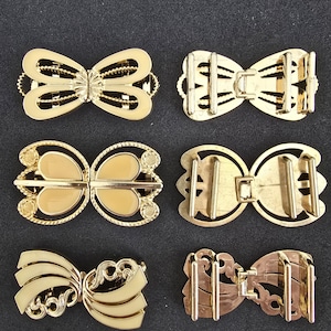 Könnte beinhalten: Sechs goldfarbene Metallverschlüsse in Schleifenform. Die Verschlüsse weisen verschiedene Designs auf, darunter filigrane Arbeiten, Emaille und dekorative Kanten. Sie sind auf dunklem Hintergrund angeordnet, um ihre komplizierten Details und ihren metallischen Glanz hervorzuheben. Diese Verschlüsse eignen sich für Kleidung oder Accessoires.
