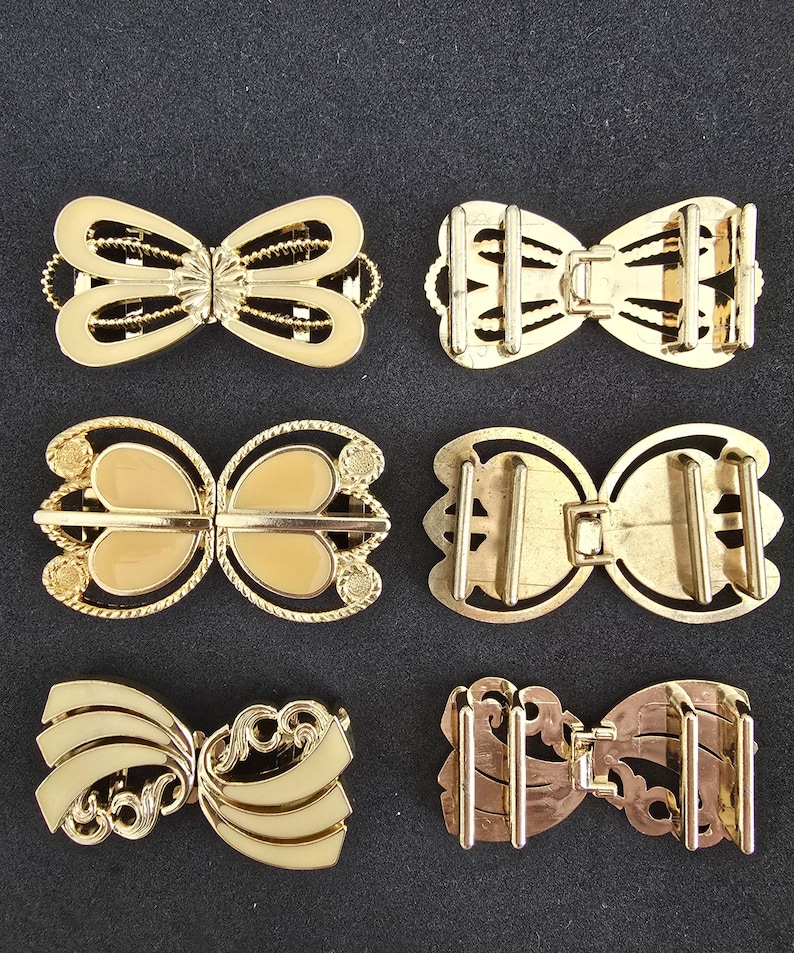 May include: Six gold-toned metal bow-shaped clasps. The clasps have various designs, including openwork, enamel, and decorative edges. The clasps are arranged on a dark background, showcasing their intricate details and metallic sheen. These clasps are suitable for clothing or accessories.