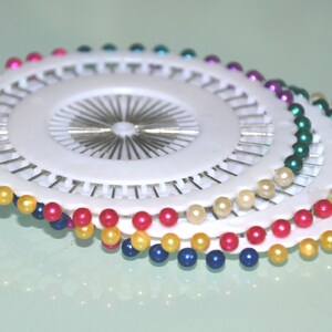 May include: A collection of white pin cushions with colorful pearl-headed pins. The pins are arranged in a circular pattern, with colors including yellow, blue, pink, green, and purple. The pins are used for sewing and crafting.