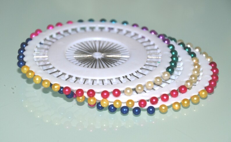 May include: A collection of white pin cushions with colorful pearl-headed pins. The pins are arranged in a circular pattern, with colors including yellow, blue, pink, green, and purple. The pins are used for sewing and crafting.