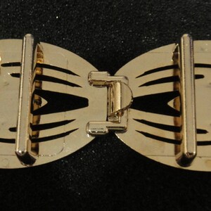 May include: A gold-colored metal bow-shaped buckle with a central clasp and two vertical bars on each side. The buckle has a decorative, openwork design with curved edges and a shiny finish. The background is black.
