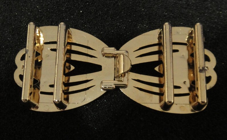 May include: A gold-colored metal bow-shaped buckle with a central clasp and two vertical bars on each side. The buckle has a decorative, openwork design with curved edges and a shiny finish. The background is black.