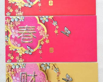 May include: Three colorful red, pink, and yellow Chinese New Year envelopes. Each envelope features gold floral designs, butterflies, and Chinese characters. The envelopes are decorated with gold accents and are likely used for gifting money during the holiday.