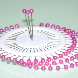May include: A white pin cushion with multiple rows of pink-headed pins arranged in a circular pattern. Several pins stand upright in the center. The pins are used for sewing and crafting.