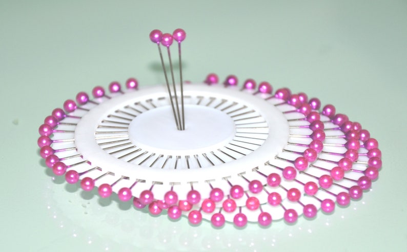May include: A white pin cushion with multiple rows of pink-headed pins arranged in a circular pattern. Several pins stand upright in the center. The pins are used for sewing and crafting.
