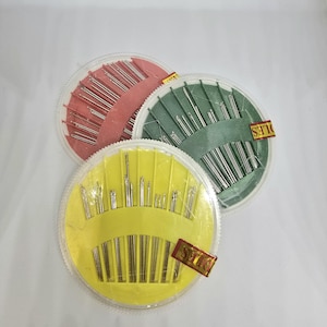 Peut inclure: Trois ensembles d'aiguilles circulaires dans des boîtiers en plastique transparent. Les boîtiers sont de couleur rose, verte et jaune. Chaque boîtier contient plusieurs aiguilles à coudre argentées. Les boîtiers portent une étiquette "LES".