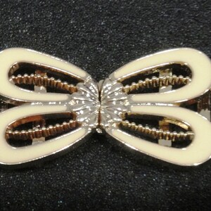 May include: A decorative metal clasp with a butterfly design. The clasp features a cream-colored enamel finish with gold-colored accents and a silver-colored center. The clasp is designed to be used on a garment or accessory.