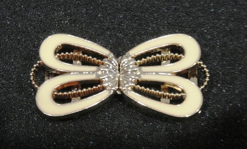 May include: A decorative metal clasp with a butterfly design. The clasp features a cream-colored enamel finish with gold-colored accents and a silver-colored center. The clasp is designed to be used on a garment or accessory.