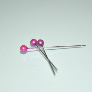 May include: Three sewing pins with metallic pink ball heads and silver shafts are arranged on a light blue surface. The pins are angled, with the points facing right. The heads have a glossy finish.