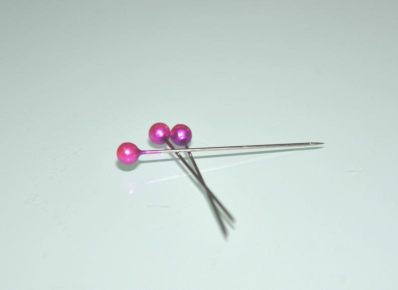 May include: Three sewing pins with metallic pink ball heads and silver shafts are arranged on a light blue surface. The pins are angled, with the points facing right. The heads have a glossy finish.