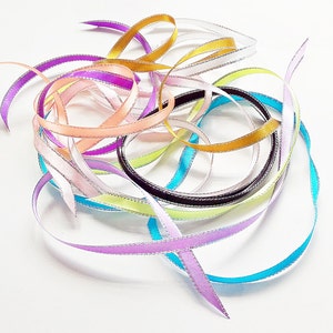 Puede incluir: Una colección de cintas coloridas en tonos morado, rosa, dorado y azul, con un borde plateado. Las cintas están entrelazadas y dispuestas sobre un fondo blanco, creando un aspecto decorativo y festivo.