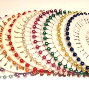 May include: A collection of white pin holders, each with a circular design and a variety of colorful pins. The pins have pearl-like heads in shades of green, purple, red, orange, white, pink, yellow, and blue. The pins are arranged in a radial pattern.