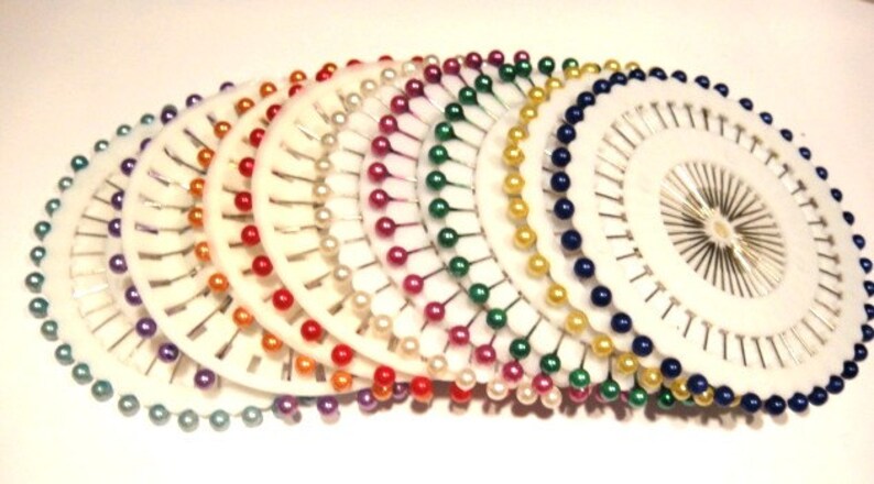 May include: A collection of white pin holders, each with a circular design and a variety of colorful pins. The pins have pearl-like heads in shades of green, purple, red, orange, white, pink, yellow, and blue. The pins are arranged in a radial pattern.