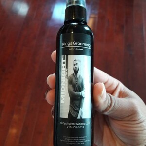 May include: A black bottle of Kings Grooming Midnight Cologne Body Spray. The bottle has a black label with white text and a photo of a man. The text on the label reads "Kings Grooming by Char's Creations", "Midnight Cologne Body Spray", "shopcharscreations.com", "215-205-1018", and "Use: Fragrance body and clothes. Shake well before spraying. Ingredients: Distilled water with various fragrance. © 2018"