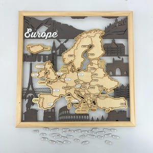 May include: Wooden Europe map with country labels, framed in light wood. The map features laser-cut details of landmarks and mountains. The word "Europe" is written in white script at the top. Includes small country name tags.
