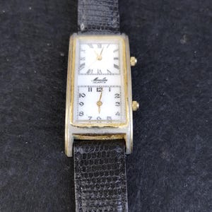 Pu&ograve; includere: Orologio rettangolare vintage con quadrante bianco e dettagli dorati. L'orologio presenta un cinturino in pelle nera e il marchio "Moulin Quartz". L'orologio ha due quadranti separati, uno con numeri romani e l'altro con numeri arabi.