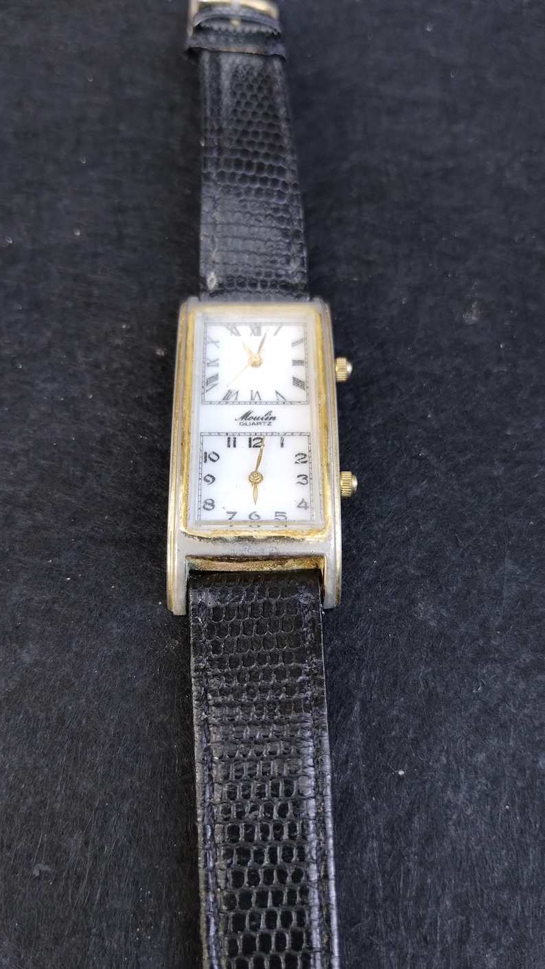 Pu&ograve; includere: Orologio rettangolare vintage con quadrante bianco e dettagli dorati. L'orologio presenta un cinturino in pelle nera e il marchio "Moulin Quartz". L'orologio ha due quadranti separati, uno con numeri romani e l'altro con numeri arabi.