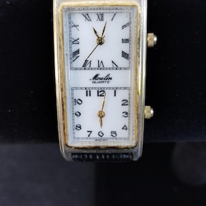 Pu&ograve; includere: Un orologio al quarzo rettangolare con quadrante bianco e numeri romani neri. L'orologio ha una cornice color oro e un cinturino in pelle nera. Il quadrante &egrave; diviso in due sezioni, con il marchio "Moulin Quartz" stampato sulla sezione superiore.